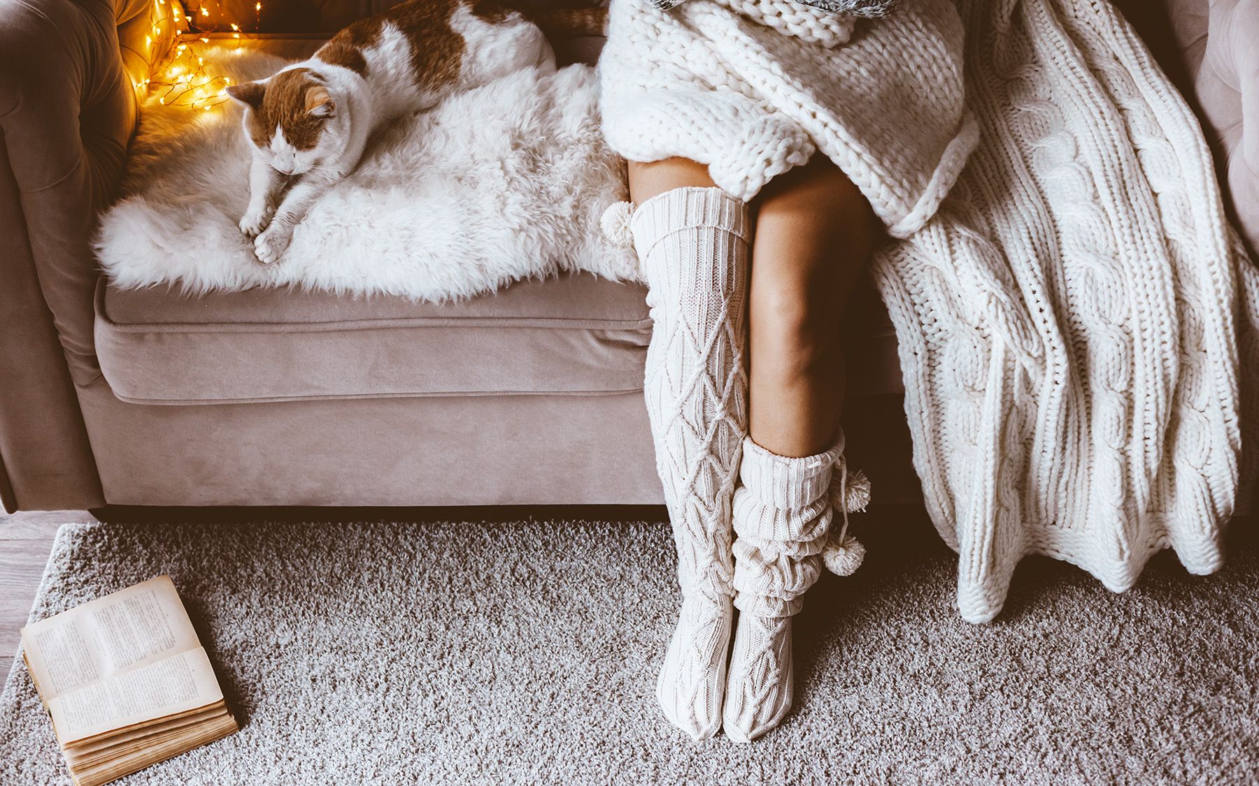 Žena v teplých ponožkách sedí na kraji postele, vedľa seba má mačku a deku, pod nohami koberec.