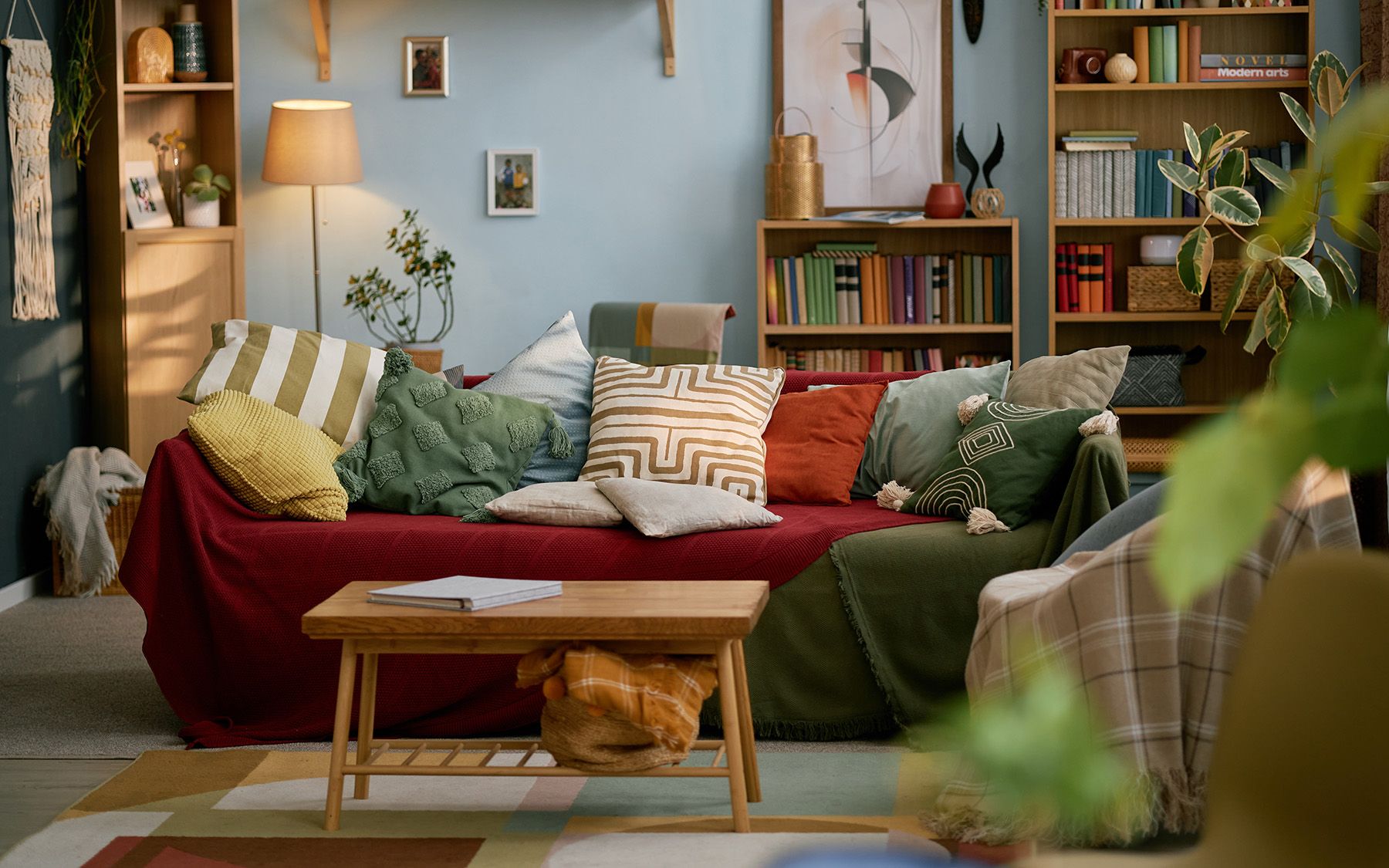 Maximalisticky zariadená obývacia izba s červeným gaučom, vankúšikmi, rastlinami, kobercom a knihami.