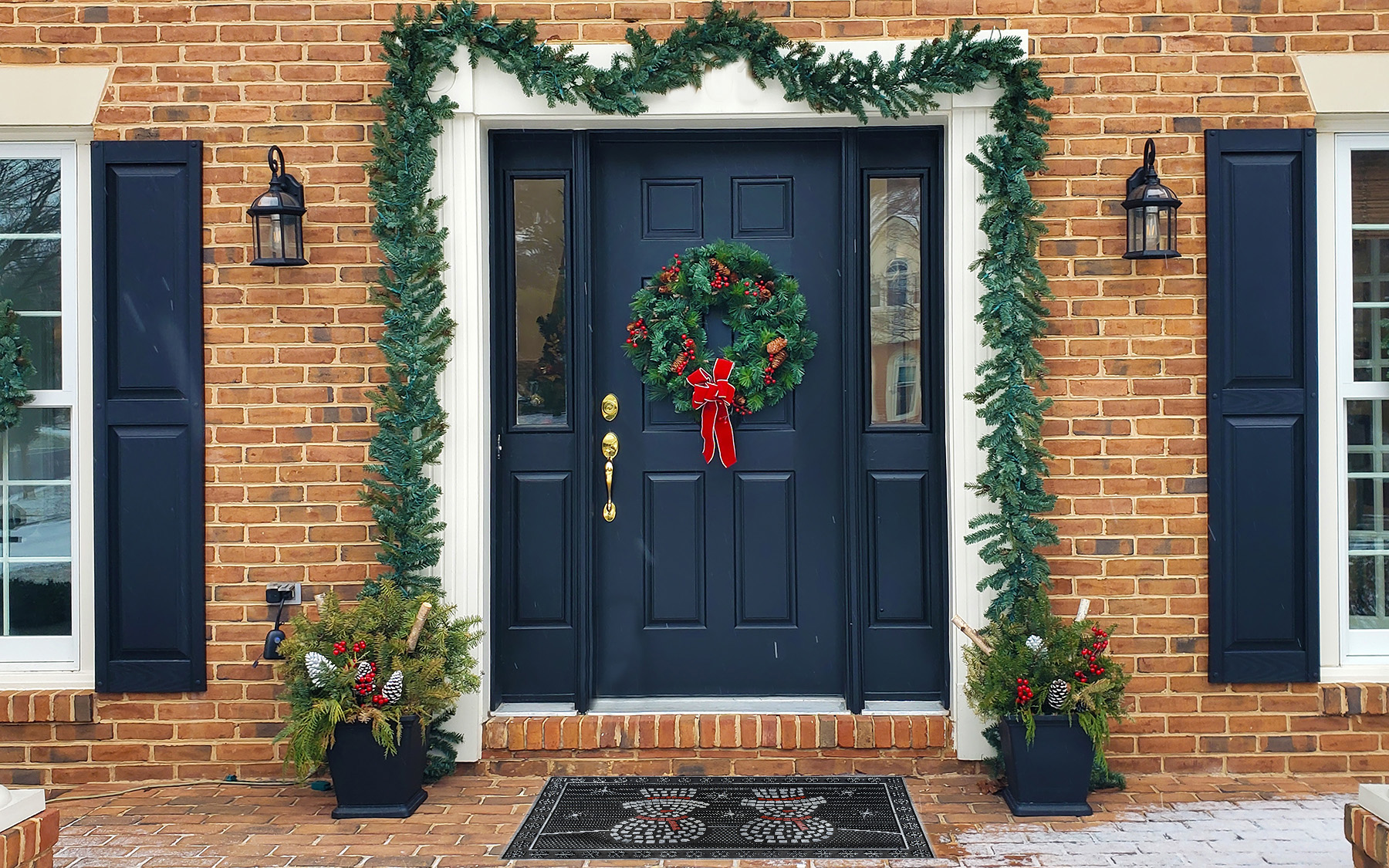 Zasnežené vchodové dvere so zimnou výzdobou a rohožkou so snehuliakmi.